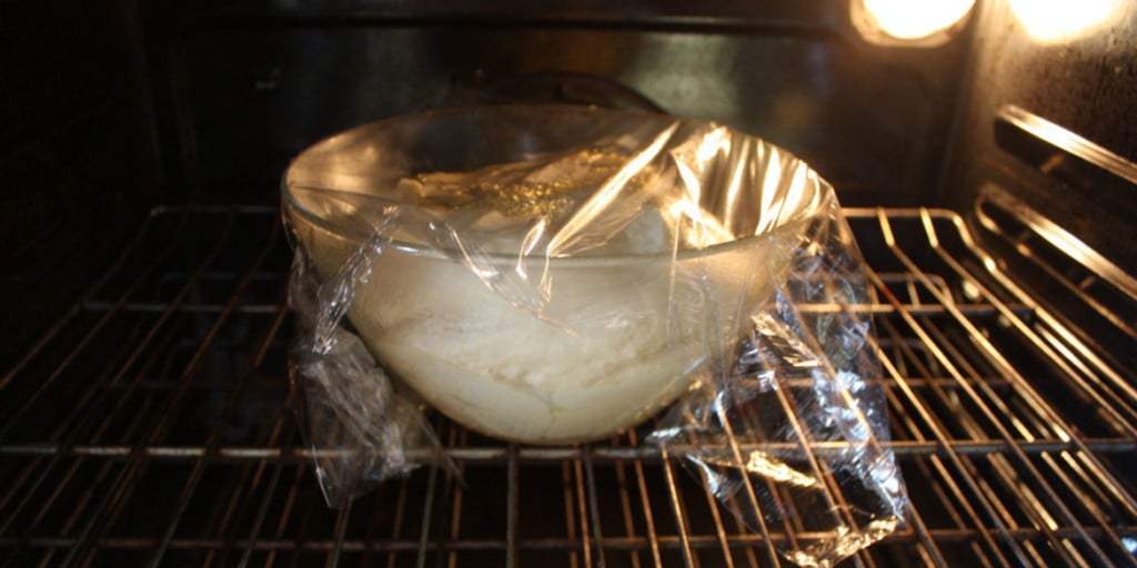 Bread dough in a glass bowl covered in oiled plastic wrap.