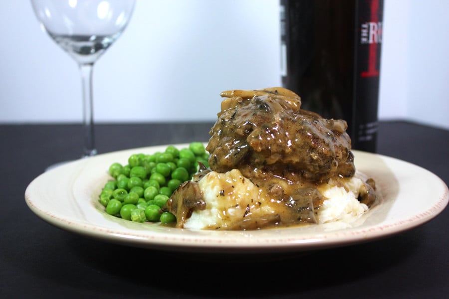 Salisbury steak on a bed of mashed potatoes with peas on a white plate.