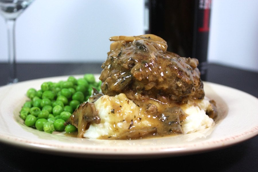 Salisbury steak covered in gravy on a bed of mashed potatoes with peas on a white plate.