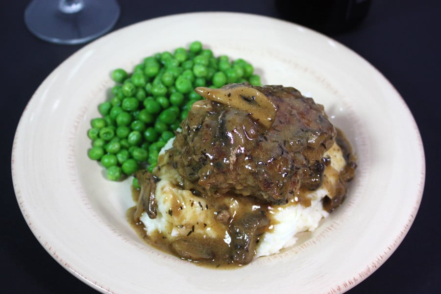 Salisbury steak covered in gravy on a bed of mashed potatoes with peas on the side.