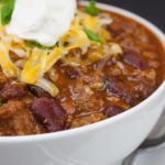 Thick and hearty homestyle chili with green onion, cheese, and sour cream as a garnish in a white bowl.
