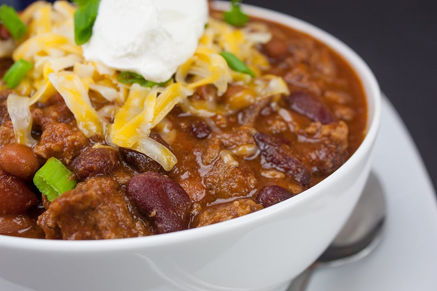 Thick and hearty homestyle chili with green onion, cheese, and sour cream as a garnish in a white bowl.