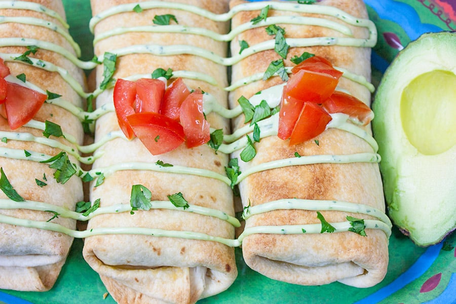 Chicken chimichangas on a green plate garnished with avocado sauce, diced tomatoes, and cilantro.