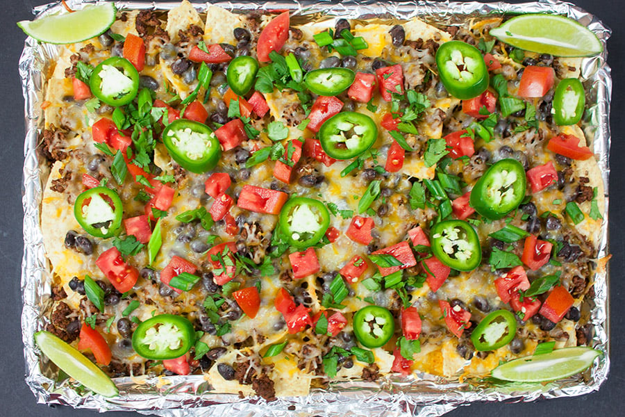 Sheet Pan Nachos topped with tomatoes, green onions, jalapenos, and lime wedges.
