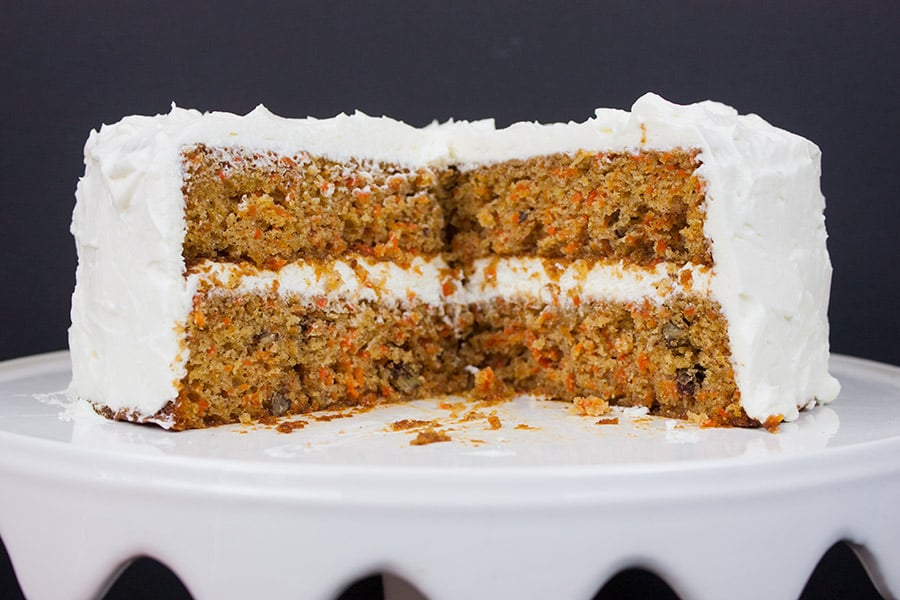 A whole homemade carrot cake cut to show inside on a white cake stand.
