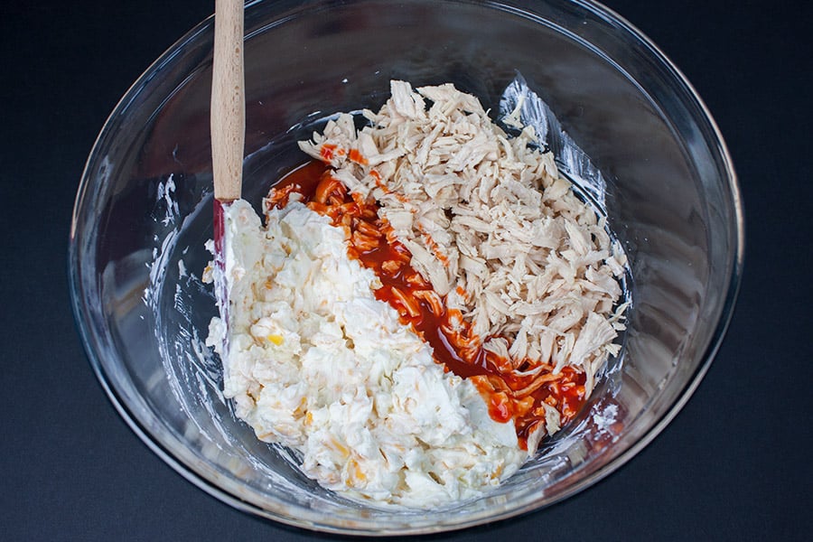 Shredded chicken, cream cheese, blue cheese, cheddar cheese, and sour cream in a glass mixing bowl.