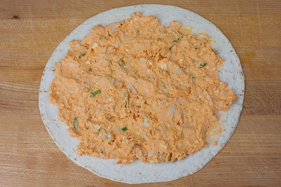 Buffalo chicken mixture spread over a flour tortilla on a cutting board.