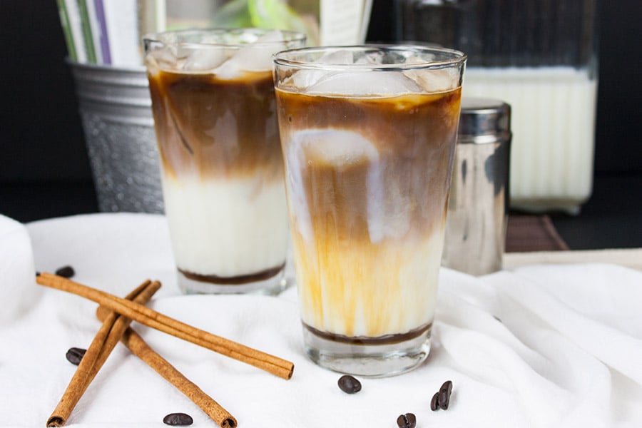 Iced Cinnamon Macchiato with cinnamon sticks and coffee beans laying on a table.