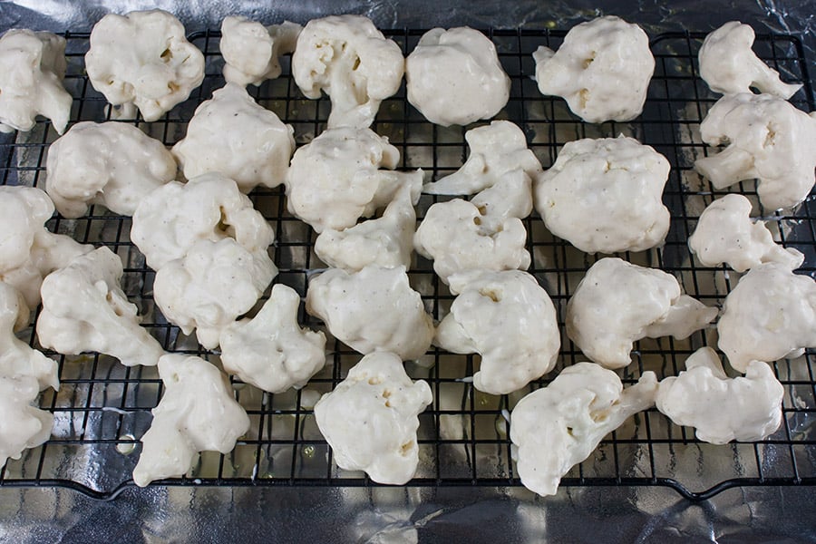 Unbaked Buffalo Cauliflower Bites dipped in batter on a wire rack in a baking sheet.