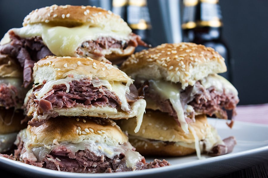 Roast beef sliders stacked on white plate beer bottles in background.