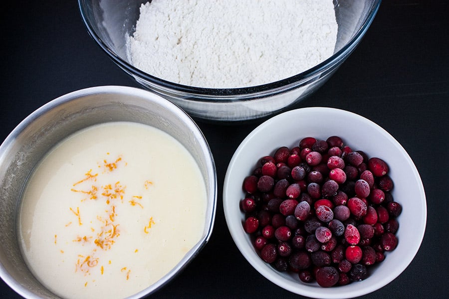 Dry flour mixture in a clear bowl, wet mixture in metal bowl, frozen cranberries in small white bowl.