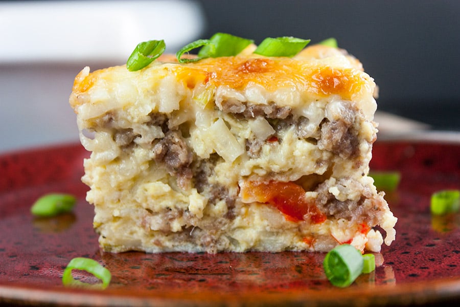 A slice of sausage hashbrown breakfast casserole on a red plate garnished with green onions.