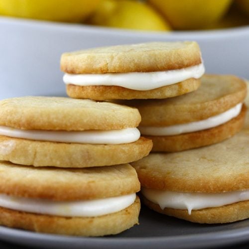 Lemon Sandwich Cookies stacked up on a plate with a bowl of lemons behind them.