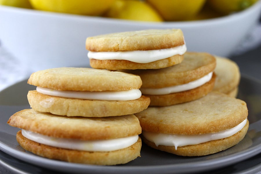 Lemon Sandwich Cookies stacked up on a plate with a bowl of lemons behind them.