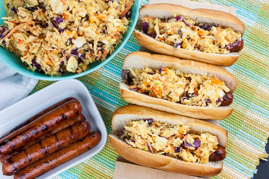 Hot Dogs topped with Mustard Slaw on a wooden hot dog board with bowl of mustard slaw in background and platter of grilled hot dogs.