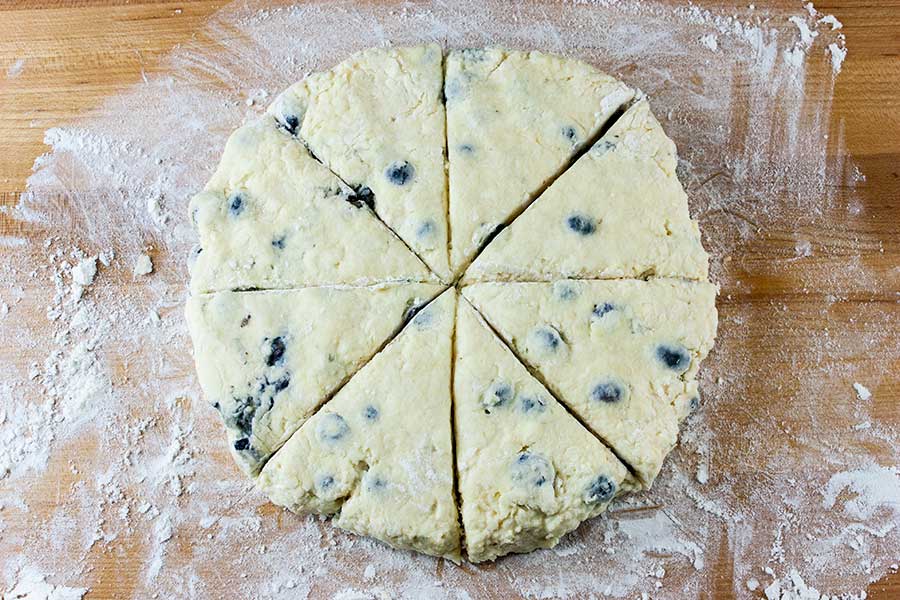 Blueberry Scone dough formed in a circle and cut into 8 pieces on a floured wooden board.