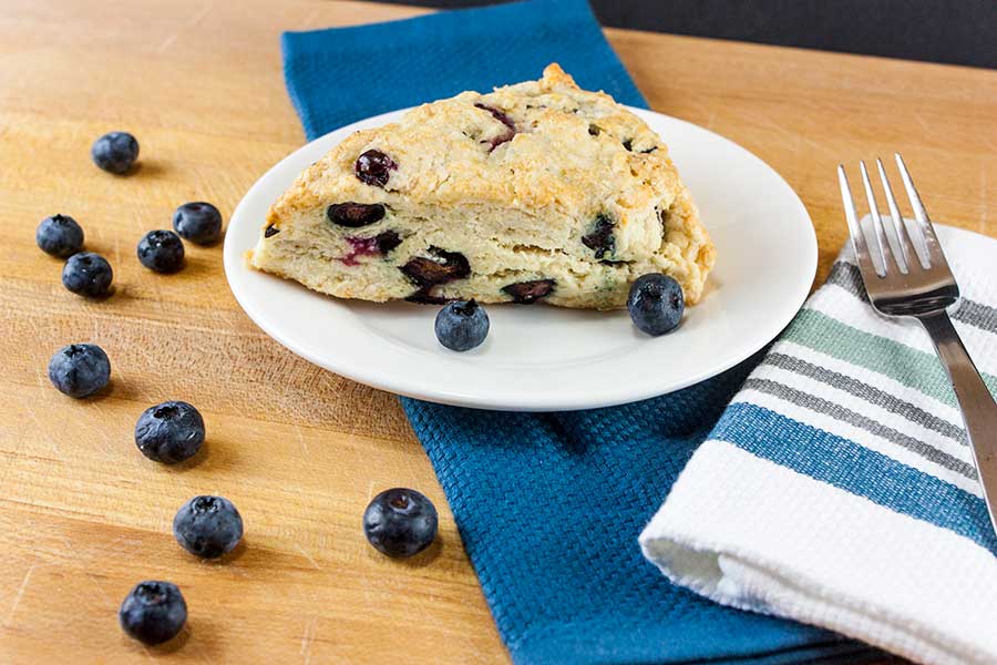 A blueberry scone on a white place with blueberries scattered across the table.