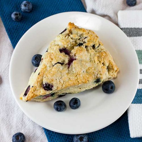 A blueberry scone on a white plate garnished with fresh blueberries.