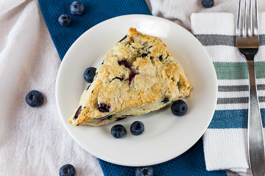 A blueberry scone on a white plate garnished with fresh blueberries.