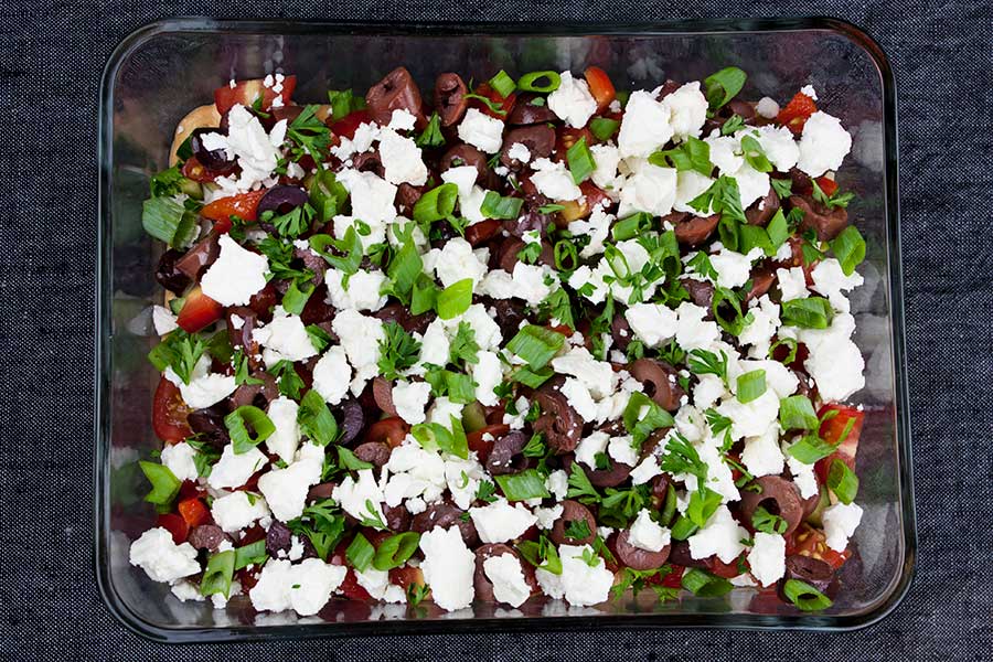 Easy Mediterranean Dip layered with hummus, sour cream, tomato, cucumber, feta cheese, Kalamata olives and green onions in a clear glass dish.