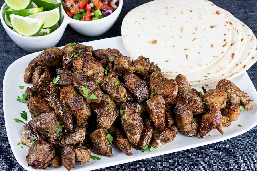 Pork carnitas on a white plate garnished with cilantro, with tortillas, lime wedges and pico de gallo.