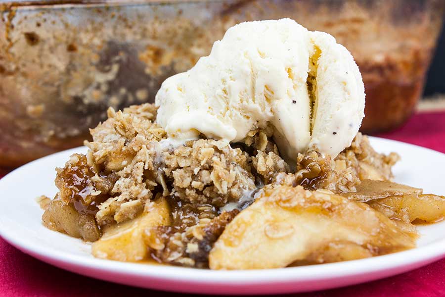A serving of the classic apple crisp topped with vanilla ice cream on a white plate.