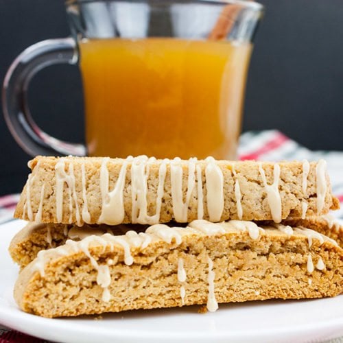 Spiced apple cider biscotti stacked on a white plate with a glass mug filled with apple cider behind them.