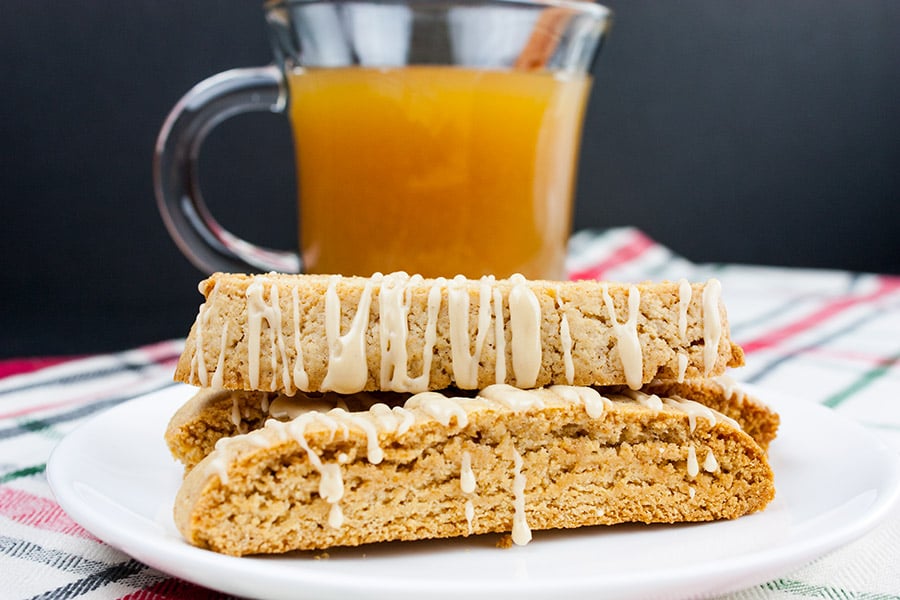 Spiced apple cider biscotti stacked on a white plate with a glass mug filled with apple cider behind them.