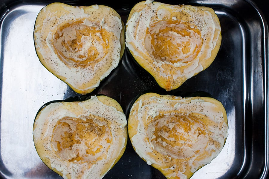 Raw acorn squash halves smeared with butter in a roasting pan.