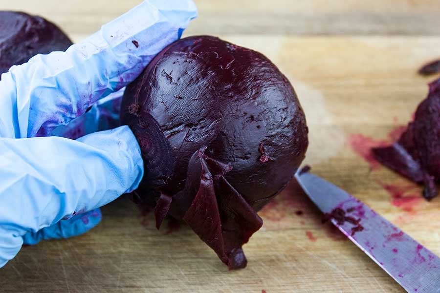 Gloved hand holding cooked beet removing the skin with a paring knife.