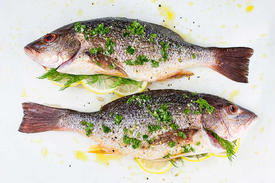 two whole red snappers prepped with herbs and seasonings on a parchment paper lined baking sheet
