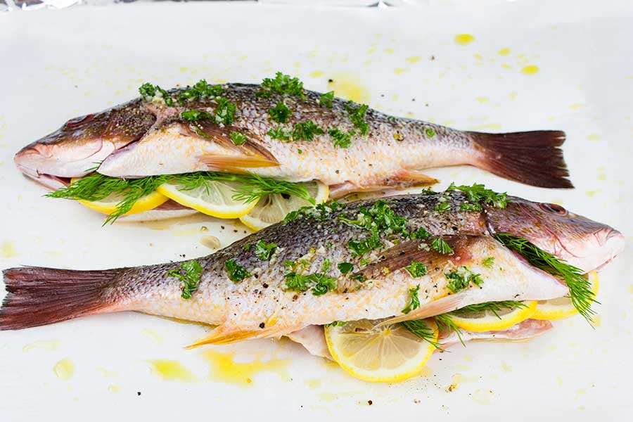 two whole red snappers prepped with herbs and seasonings on a parchment paper lined baking sheet
