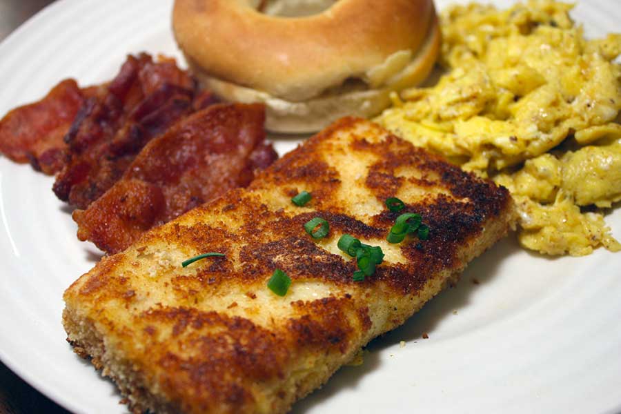 Fried Grit Cakes on a white plate with bacon, scrambles eggs, and a bagl