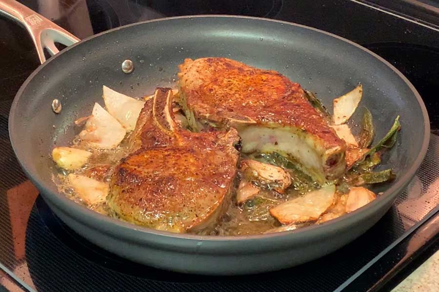 Pork chops cooking in a skillet with onion, garlic, and sage