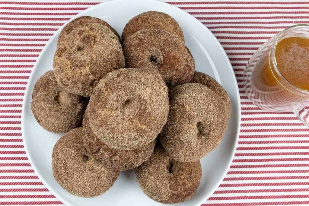 Apple cider donuts on a white platter with a cup of apple cider on the side.