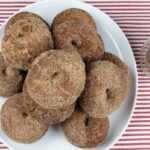 Apple cider donuts on a white platter with a cup of apple cider on the side.