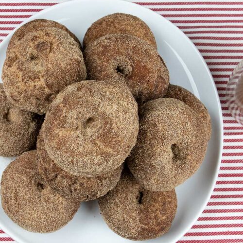 Apple cider donuts on a white platter with a cup of apple cider on the side.