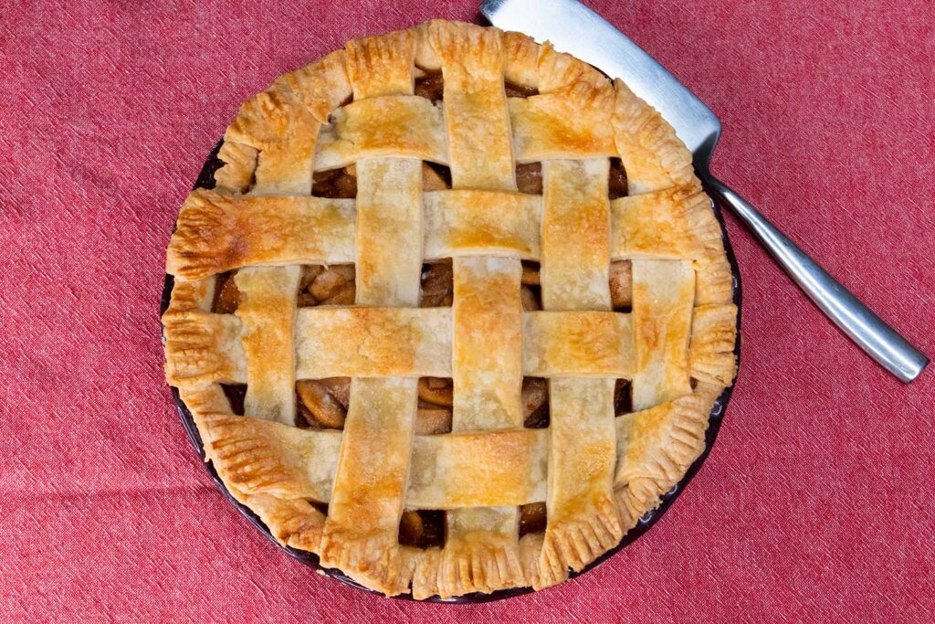 An apple pie with a lattice crust on a red table cloth with a pie server to the side.