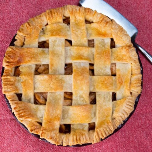 Whole apple pie on a red table cloth with a pie server to the side.