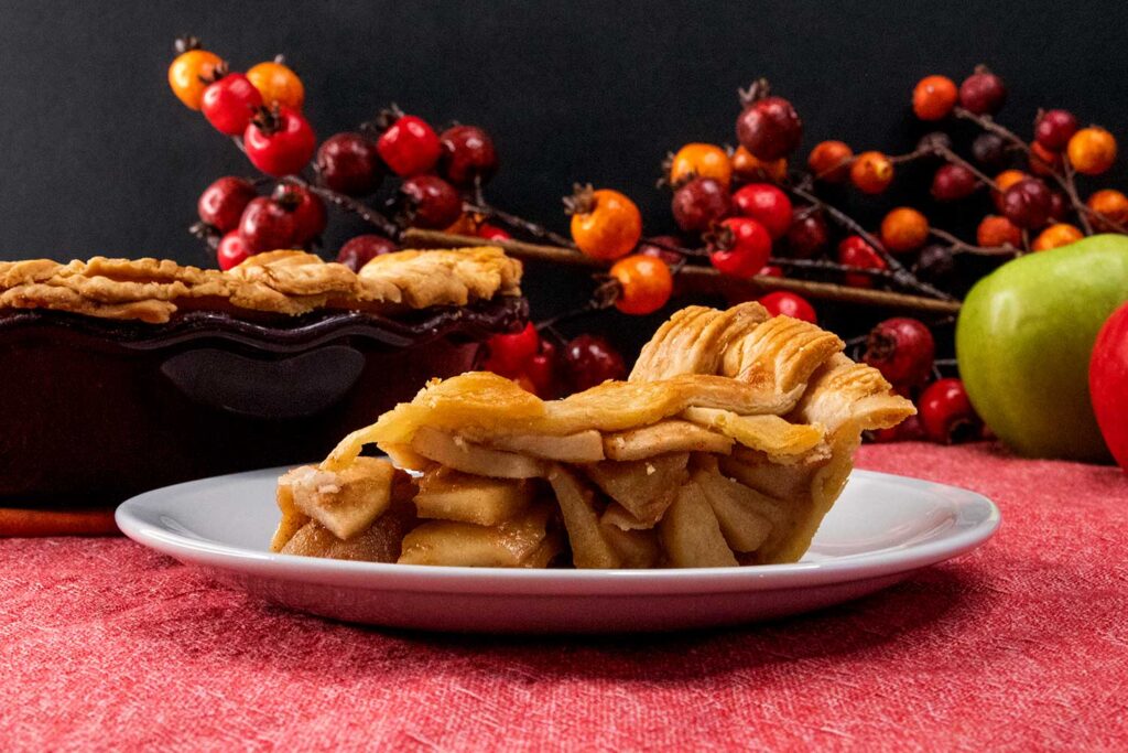 A slice of apple pie on a white plate with apples in the background.