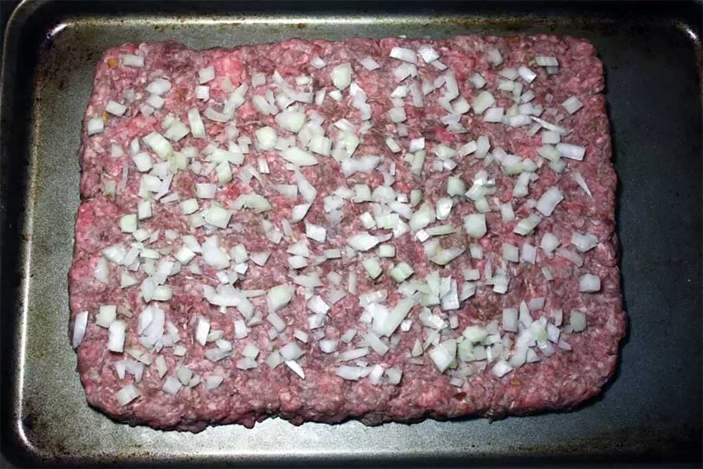 hamburger meat patted into a square on a baking tray topped with chopped onions