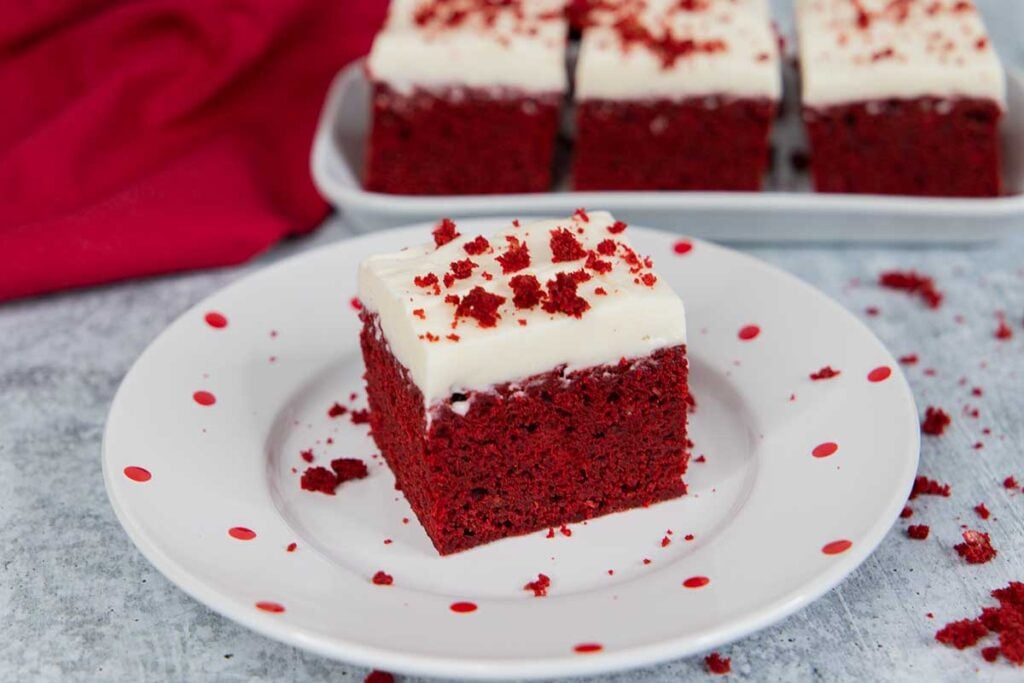 Red velvet brownie square with cream cheese frosting, garnished with brownie crumbs on a light plate with red dots.
