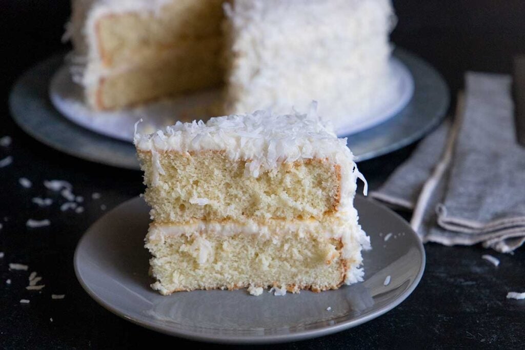A slice of coconut cake on a gray plate with the whole cake in the background.