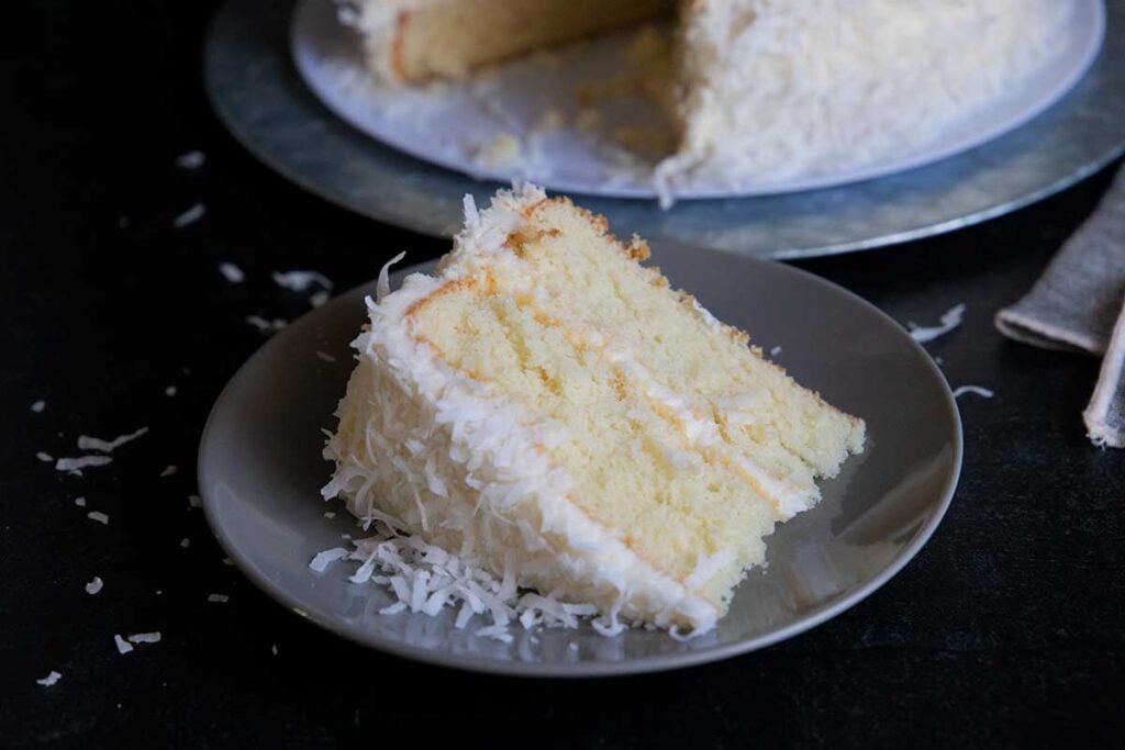 Slice of coconut cake on a light plate with the whole cake in the background.