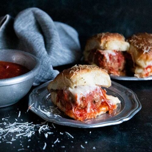 Meatball sliders on a silver plate garnished with shaved parmesan cheese.