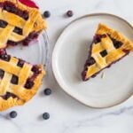 Blueberry pie slice on a white plate with the rest of the pie off to the side.