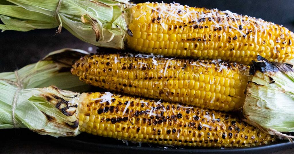 Grilled corn on the cob, topped with shredded parmesan cheese on a black plate.