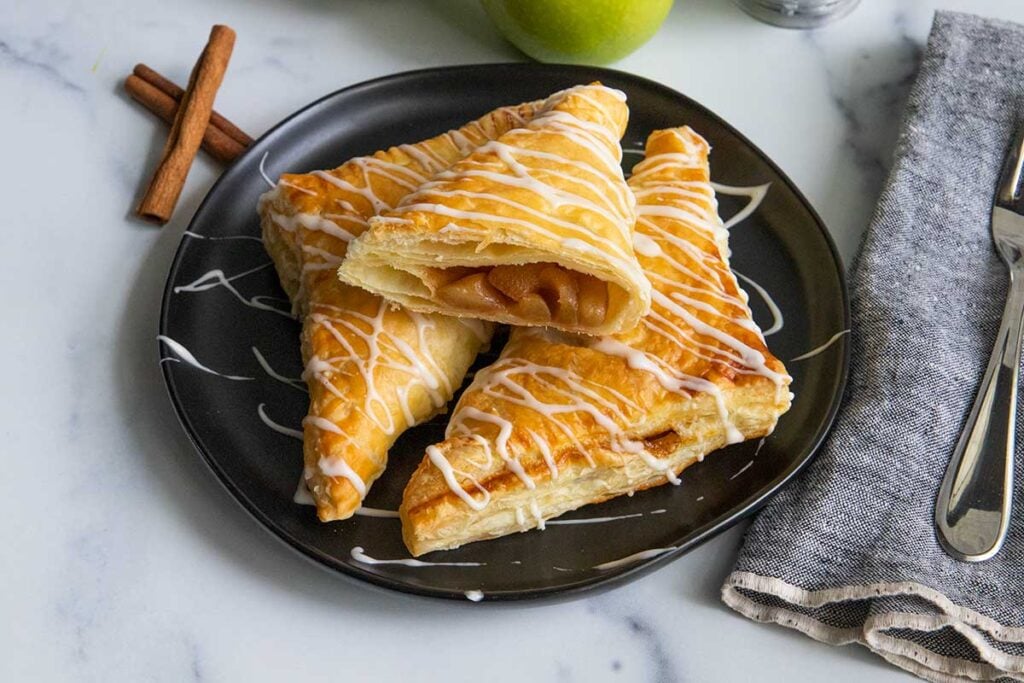 Puff pastry apple turnovers on a black plate with cinnamon sticks and apples as garnish.