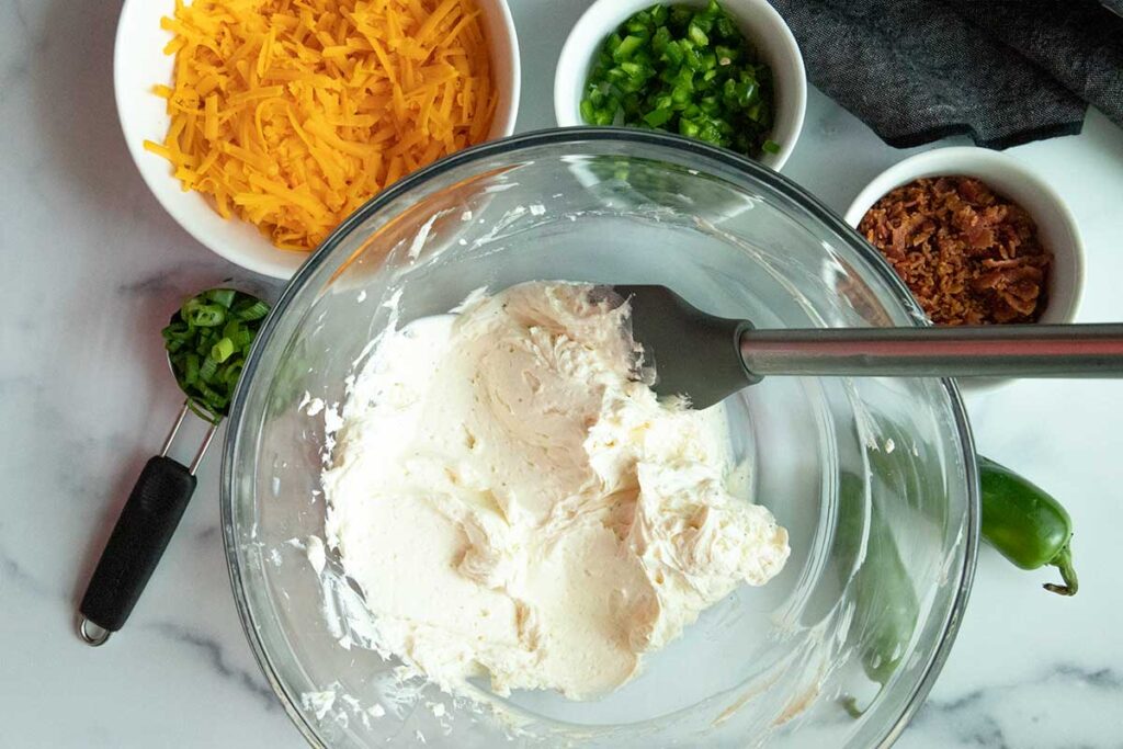 Softened cream cheese in a glass bowl surrounded by shredded cheddar cheese, chopped jalapenos, diced green onions, and chopped bacon.