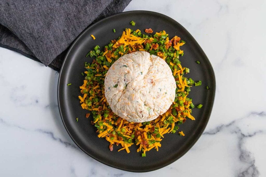 Jalapeno popper cheese ball lying on top of the toppings on a dark plate.